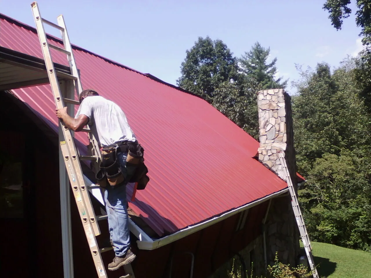 Roofer on a ladder beside a red metal roof and stone chimney — Soffit & Fascia Repair by Roofing Brothers in Sallisaw